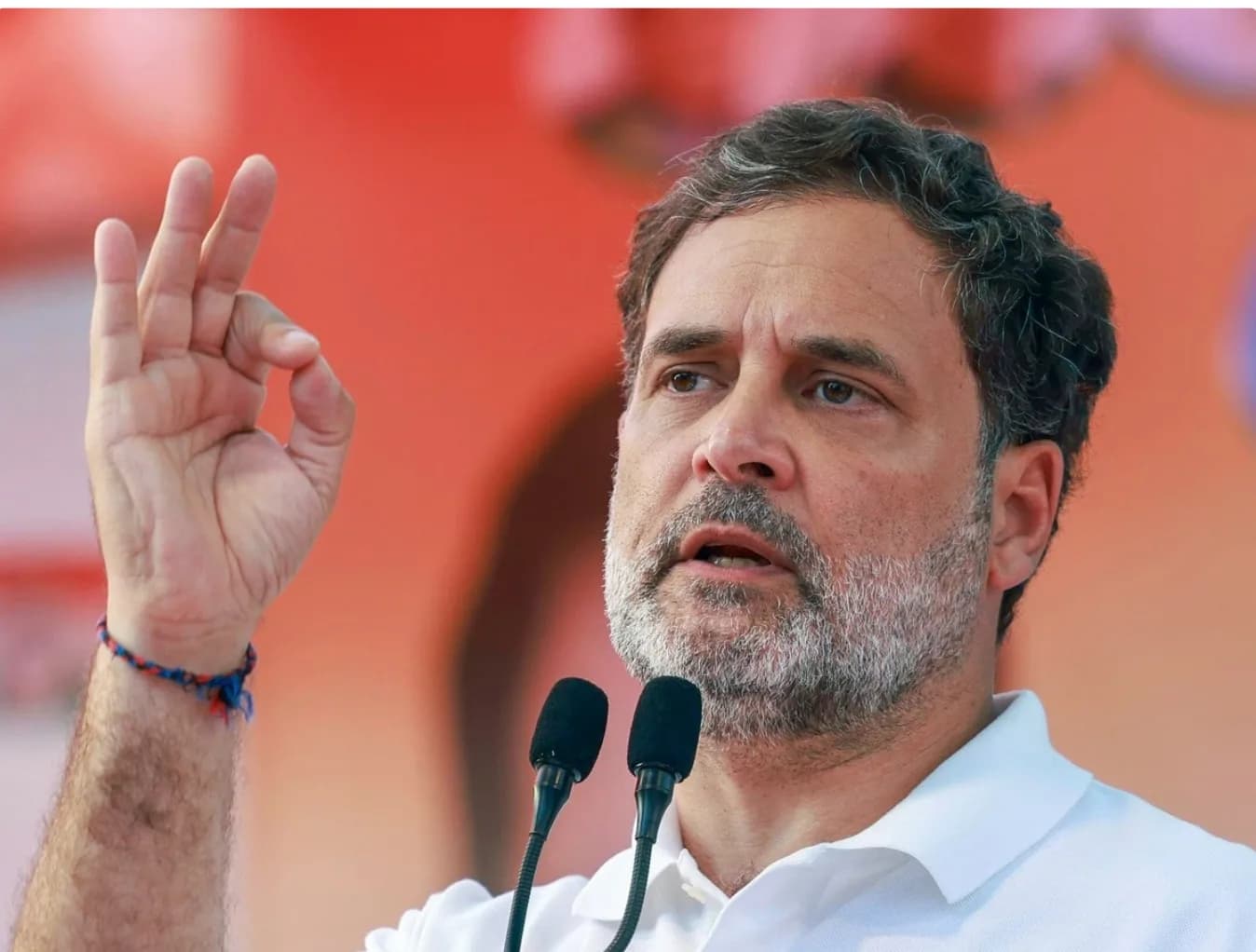 राहुल गांधी एक सफेद पोलो शर्ट पहने हुए भाषण दे रहे हैं और 'ओके' का इशारा कर रहे हैं।