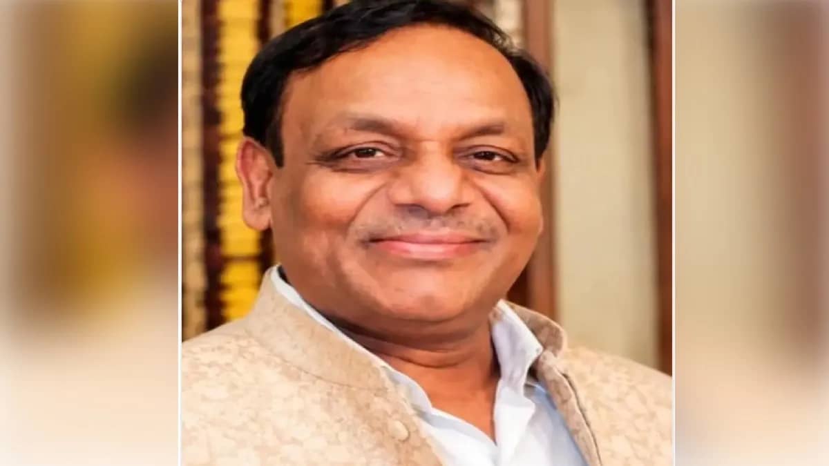 एक भारतीय व्यक्ति मुस्कुराते हुए, हल्के रंग की जैकेट और सफेद शर्ट पहने हुए।