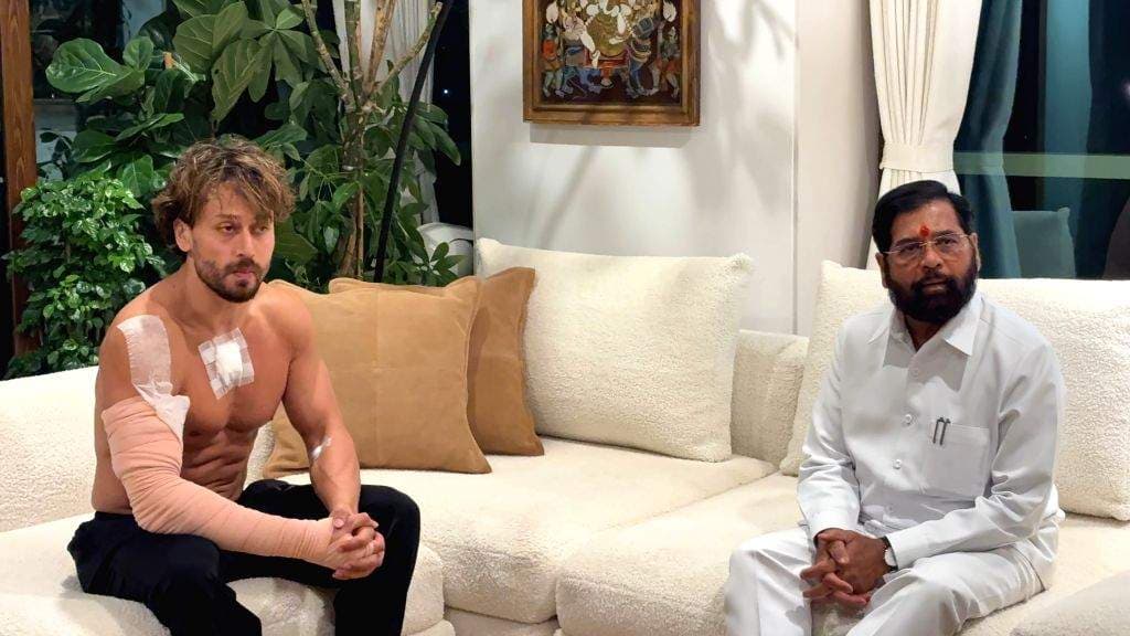 एक आदमी सफेद ब्लेज़र और काले पैंट पहने हुए लाल कालीन पर खड़ा है।