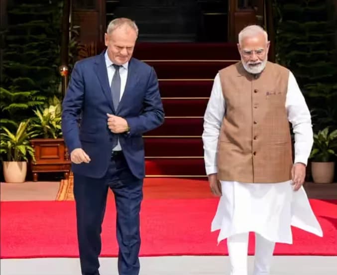 एक व्यक्ति नीले सूट में और दूसरा व्यक्ति भूरे रंग की जैकेट और सफेद कुर्ते में लाल कालीन पर चल रहे हैं।
