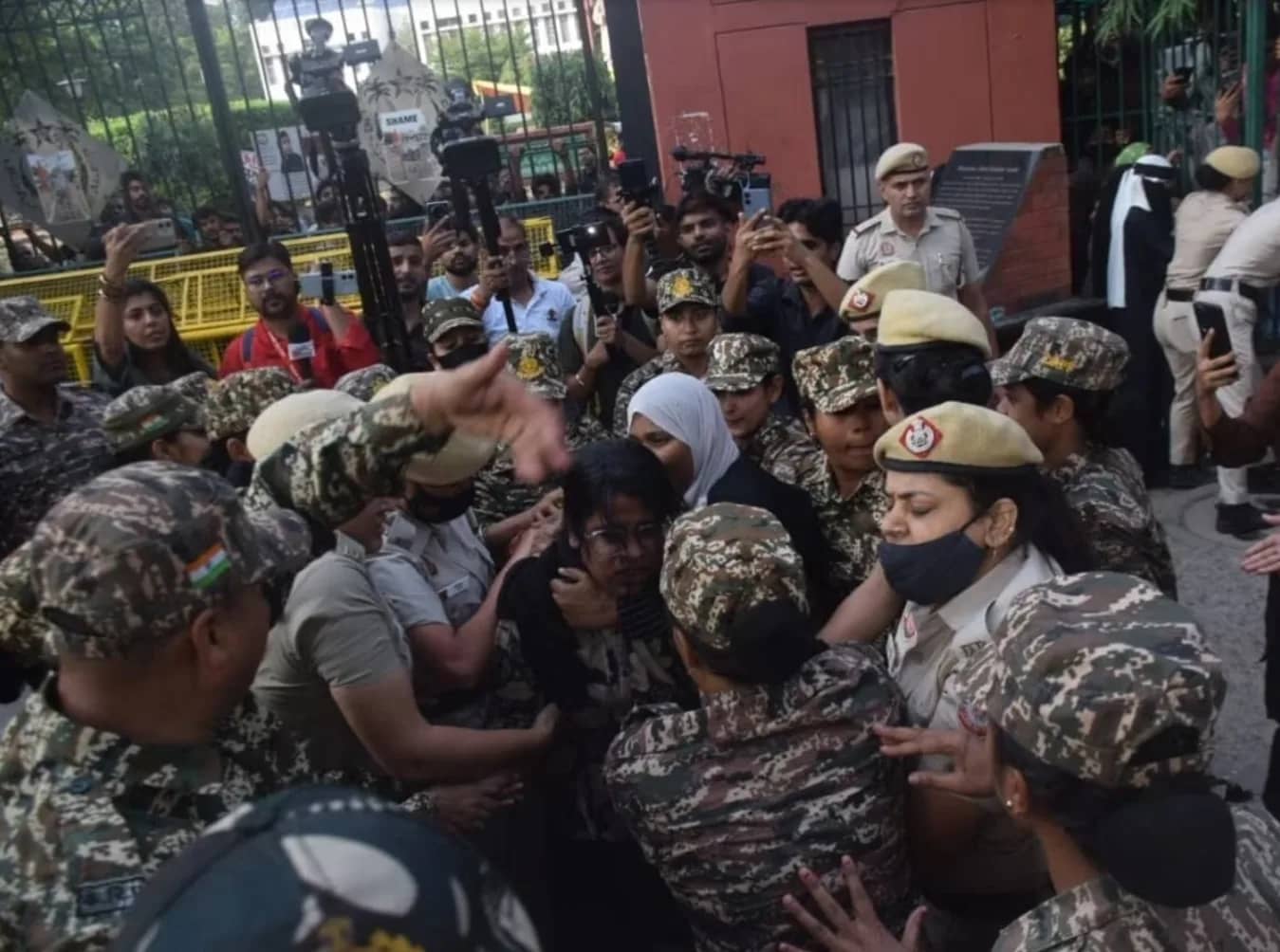 पुलिस अधिकारियों और सैन्य वर्दी पहने लोगों की भीड़ एक महिला को घेरे हुए है।
