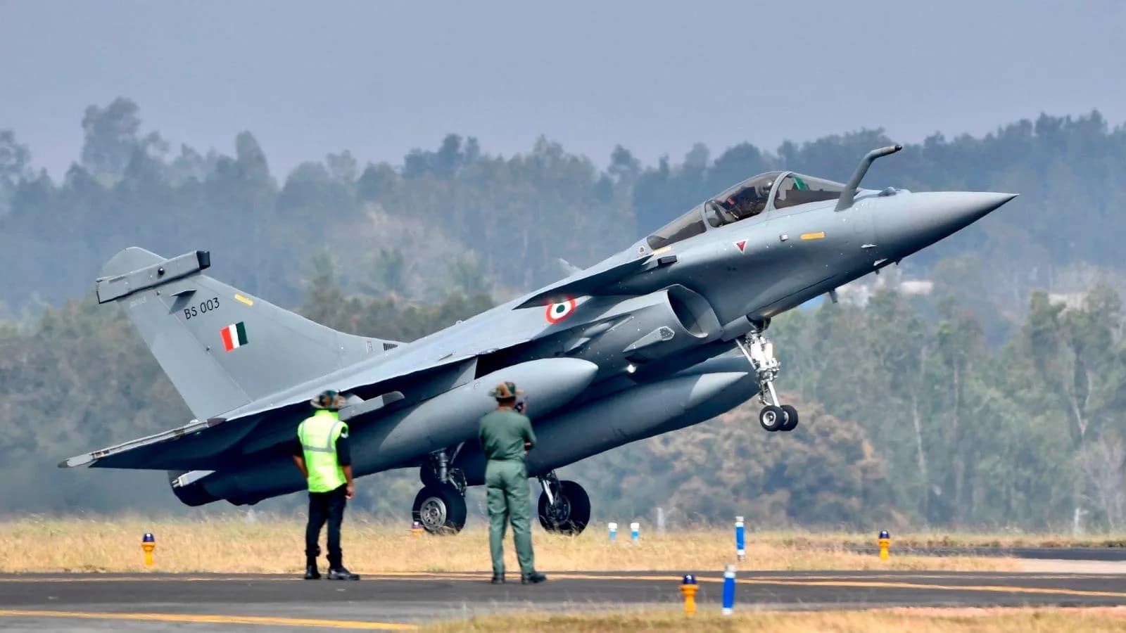 एक ग्रे फाइटर जेट रनवे से उड़ान भर रहा है, जिसके पास दो लोग खड़े हैं।