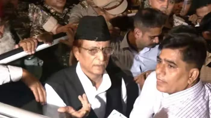 एक आदमी काले रंग की टोपी और चश्मा पहने हुए, सफेद शर्ट और काले कोट में, बात कर रहा है।