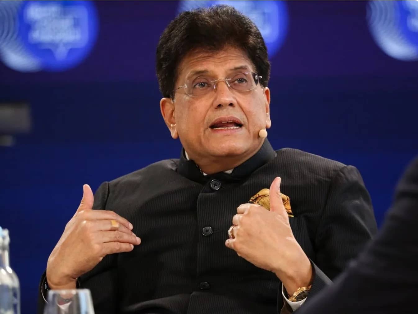 एक व्यक्ति काले रंग की शेरवानी और चश्मा पहने हुए, अपने हाथों से इशारा करते हुए बोल रहा है।