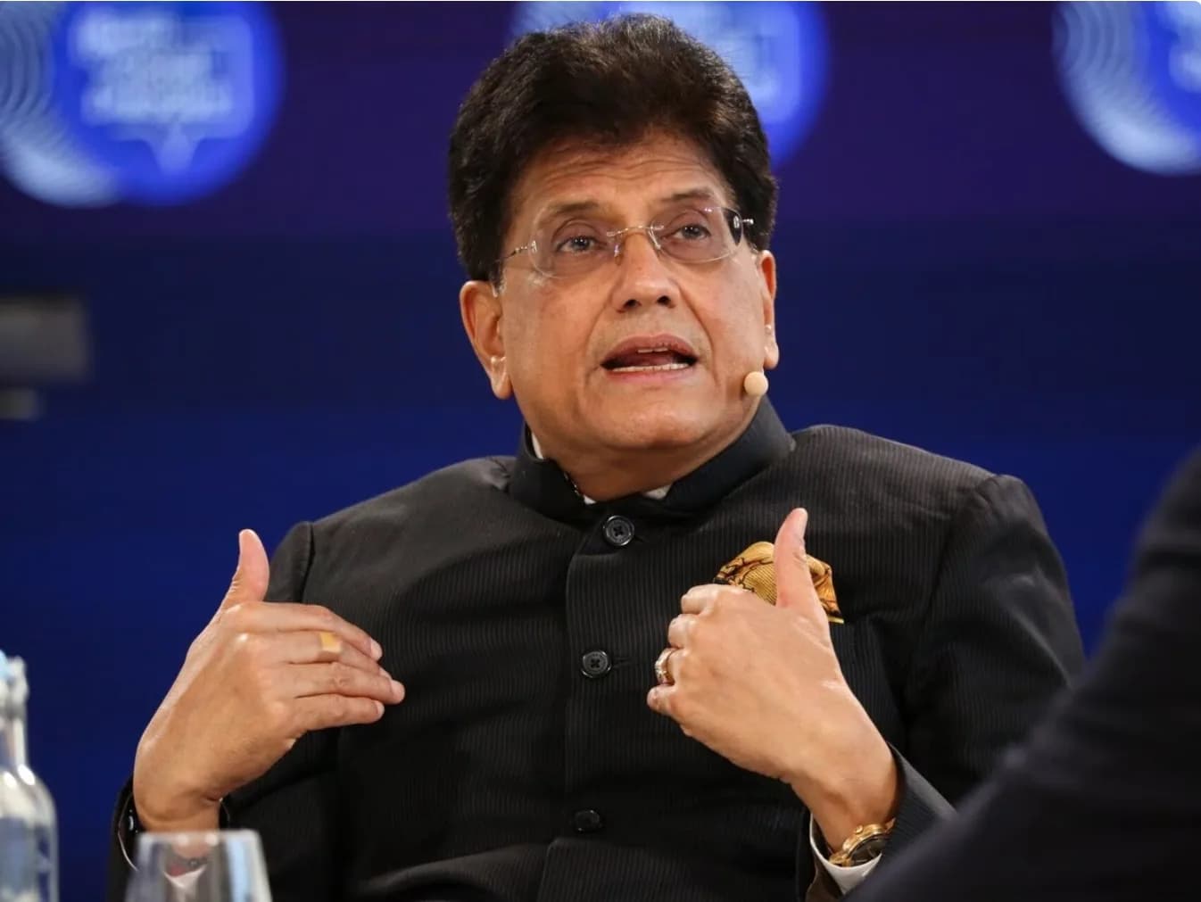 एक व्यक्ति काले रंग की शेरवानी और चश्मा पहने हुए, अपने हाथों से इशारा करते हुए बोल रहा है।