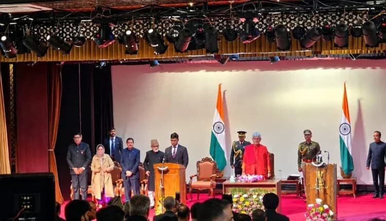 एक मंच पर भारतीय झंडों के बीच कई लोग खड़े हैं, जिनमें से एक व्यक्ति पोडियम पर बोल रहा है।