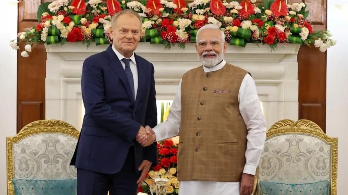 दो पुरुष एक-दूसरे से हाथ मिलाते हुए, उनके पीछे फूलों की सजावट है।