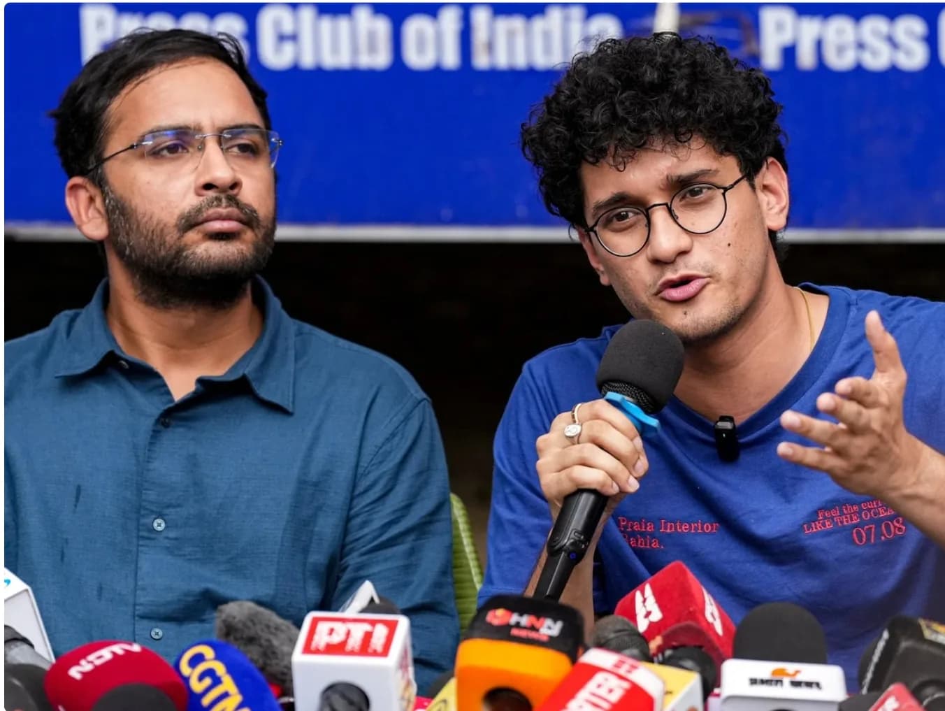 दो आदमी प्रेस कॉन्फ्रेंस में माइक्रोफोन के सामने बैठे हैं।