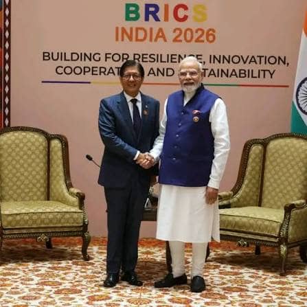 एक व्यक्ति सफेद शर्ट पहने हुए मुस्कुरा रहा है, जिसके पीछे एक लकड़ी की दीवार है।